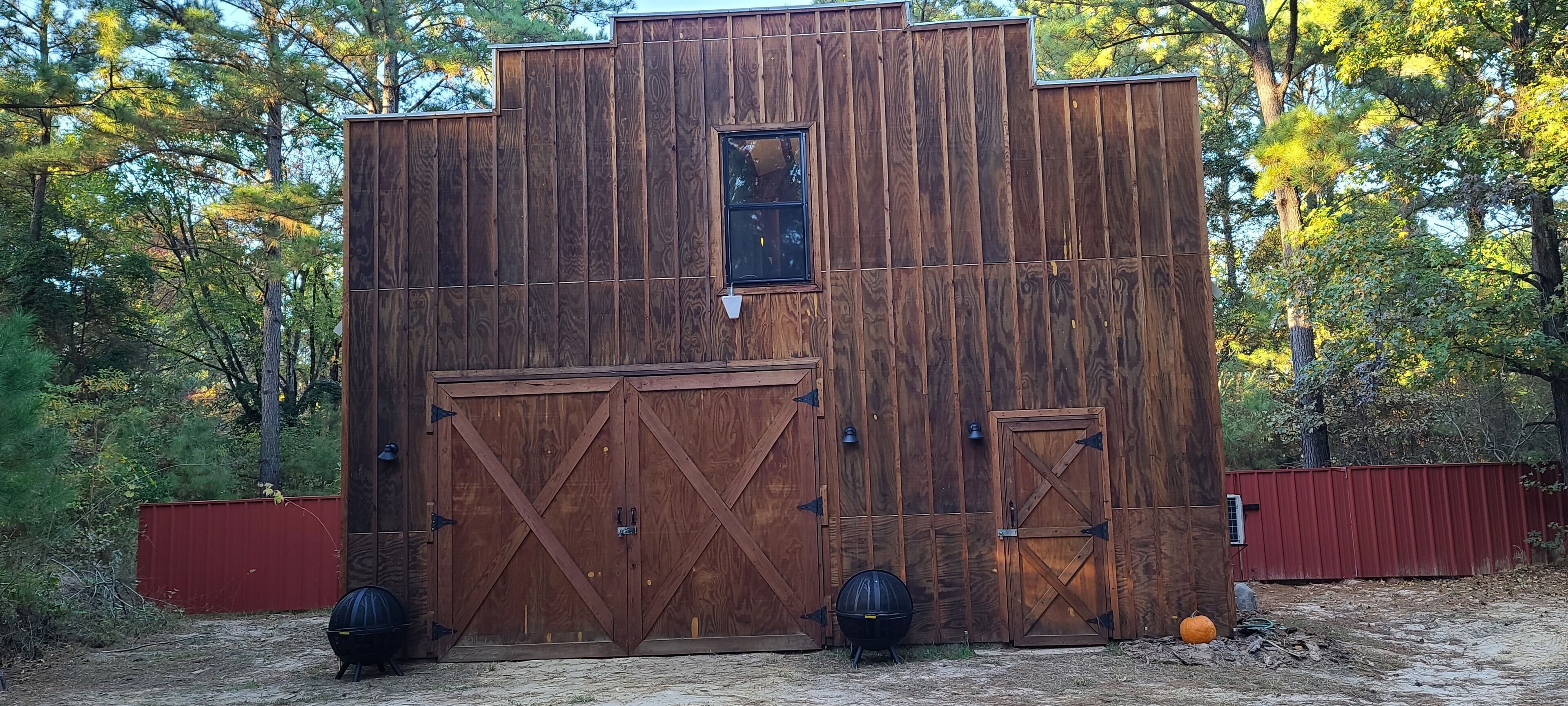 A rustic wooden structure with two large doors and a small window, surrounded by trees.