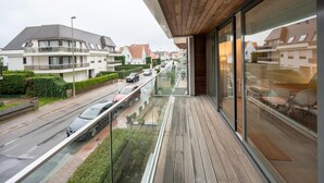 A modern balcony with a wooden floor and glass railings overlooking a street with parked cars and residential buildings. - Apartment in Knokke Near the Beach & Casino (Knokke-Heist)