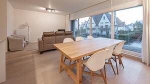 A modern living room with a wooden dining table, white chairs, a beige sofa, and a large window with a view of houses. - Apartment in Knokke Near the Beach & Casino (Knokke-Heist)