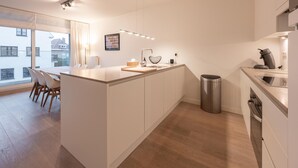 A modern kitchen with a central island, a dining area with chairs, a trash bin, and a view of the outside. - Apartment in Knokke Near the Beach & Casino (Knokke-Heist)