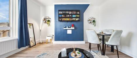 Una sala de estar moderna con una pared azul de acento, un televisor de pantalla plana, un comedor con mesa y sillas, y un gran ventanal con cortinas.
