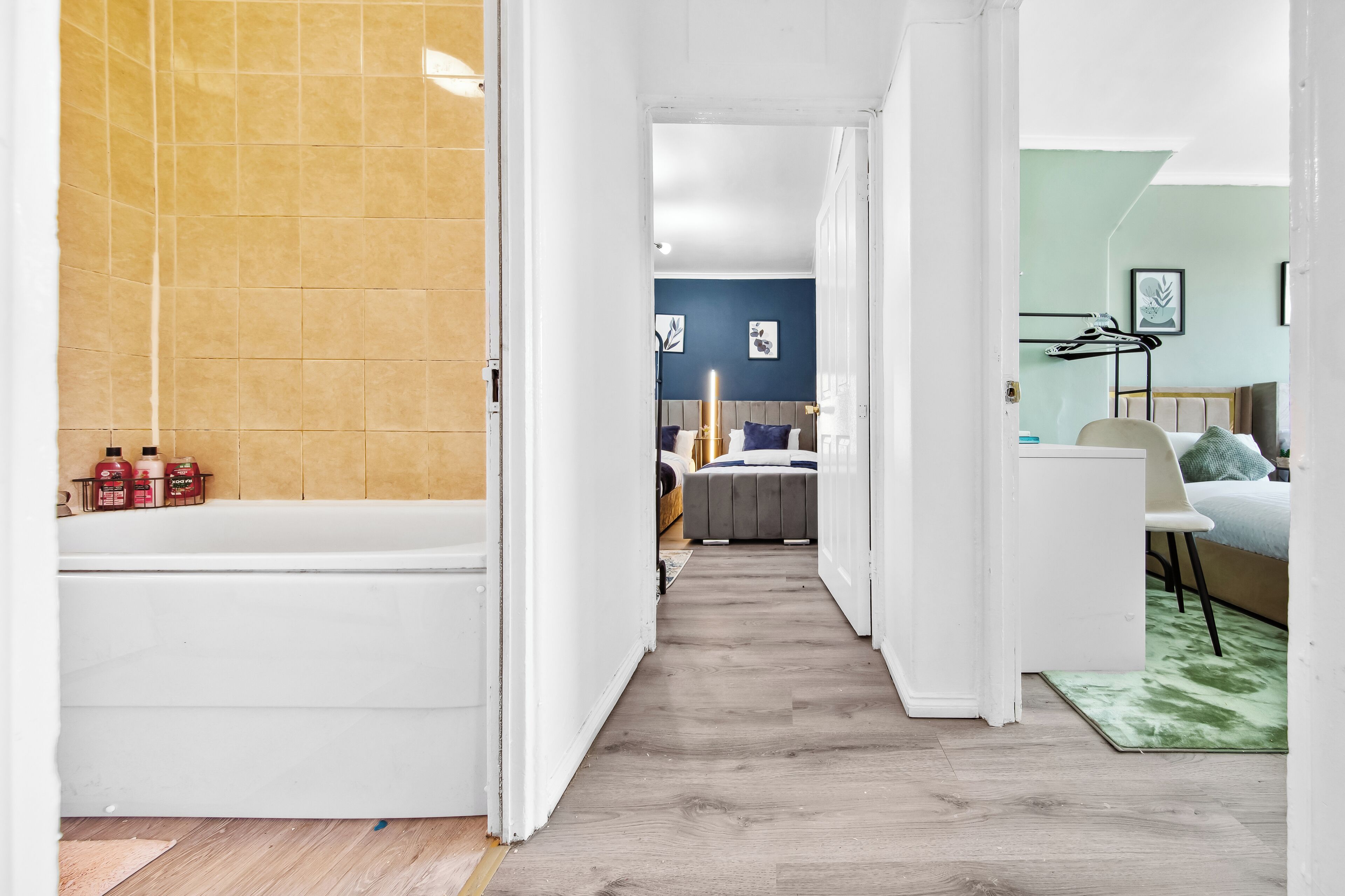 Un pasillo estrecho que conduce a un dormitorio con cama, una silla y una mesita. Se ve una bañera a la izquierda, y una alfombra verde en el suelo.