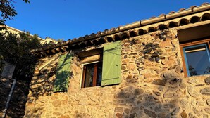 A stone building with a tiled roof, a visible window with green shutters, and a small entrance. - House in a charming village in the countryside, near the Olivettes Dam (Vailhan)
