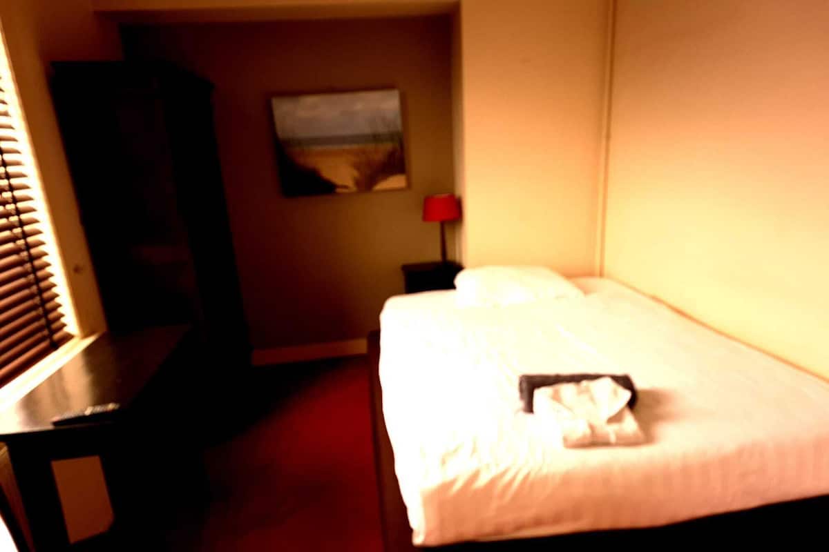 Quarto de hotel com cama, escrivaninha, cadeira, pintura na parede e abajur vermelho.