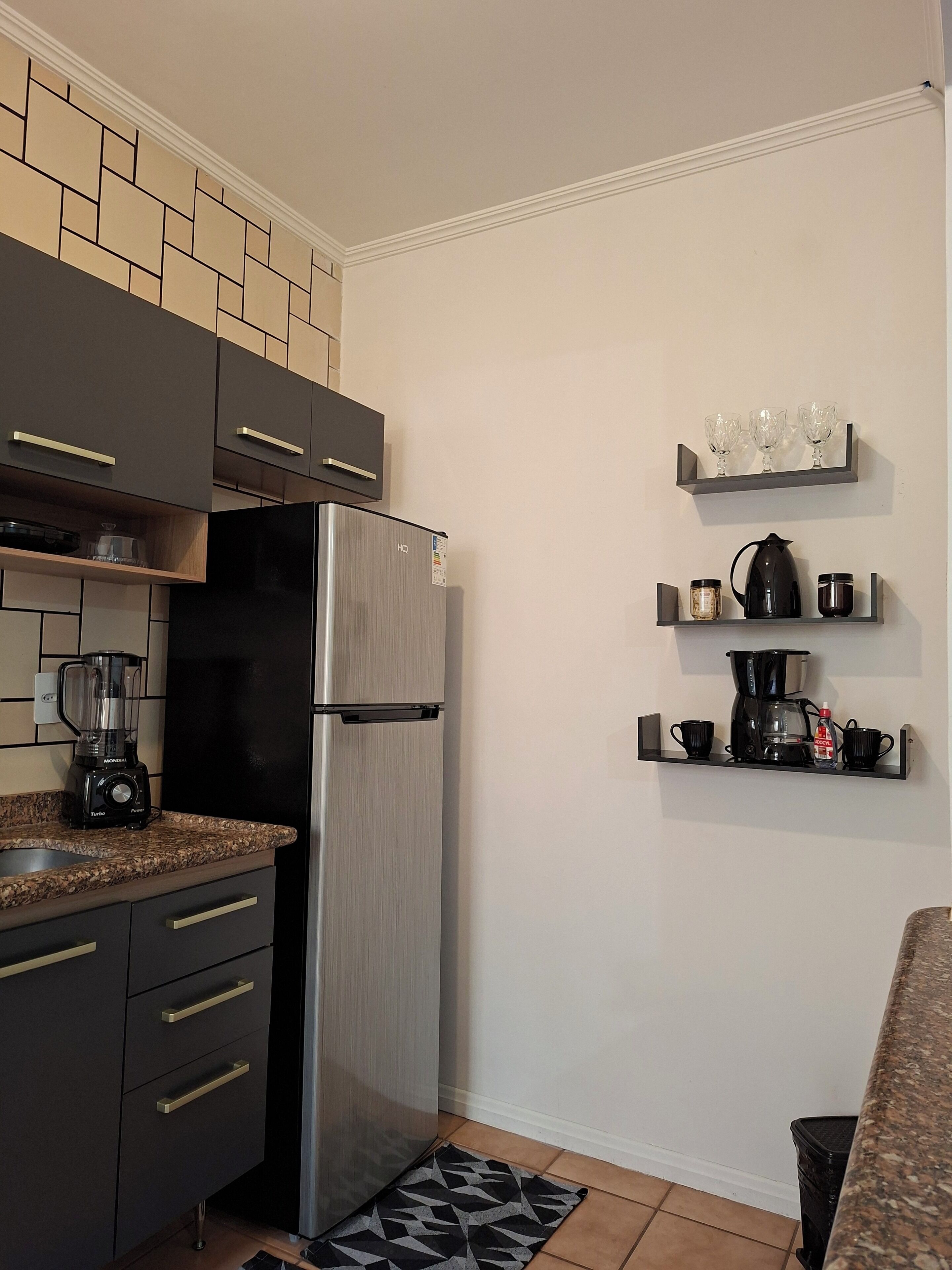 Una cocina compacta con un refrigerador de acero inoxidable, armarios oscuros, una cafetera integrada y una licuadora sobre la encimera.