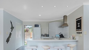 A modern kitchen with white cabinets, a central island with stools, a microwave, and a stainless steel range hood. - Flaxmill Haven - Cooks Beach Holiday Home (Whitianga)