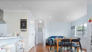 A modern kitchen with a dining area, featuring a wooden table and chairs, a blue sofa, and a kitchen counter with bar stools. - Flaxmill Haven - Cooks Beach Holiday Home (Whitianga)