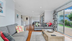 A modern living room with a grey sofa, a wooden coffee table, and a large sliding glass door leading to a balcony. - Flaxmill Haven - Cooks Beach Holiday Home (Whitianga)