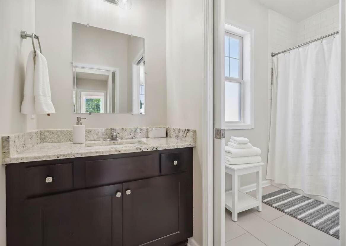Un baño moderno con encimera de mármol, gabinetes oscuros, un lavabo blanco y una zona de ducha con cortina blanca.