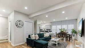 Una sala de estar moderna con un sofá seccional verde oscuro, un comedor con mesa y sillas blancas, y una cocina al fondo.
