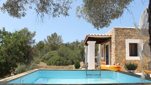 A swimming pool with a wooden deck, a stone building with a tiled roof, and surrounding greenery. - New! Villa Can Cardona (Santa Agnès de Corona)