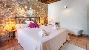 A bedroom with a stone wall, a wooden ceiling, a bed with white linens and pink pillows, two bedside lamps, and a bench against the wall. - New! Villa Can Cardona (Santa Agnès de Corona)