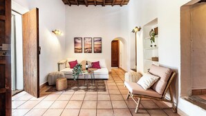 A cozy living room with a white sofa, a wooden coffee table, and a wicker ottoman. There are three framed pictures on the wall, a wooden door, and a shelf with plants. - New! Villa Can Cardona (Santa Agnès de Corona)