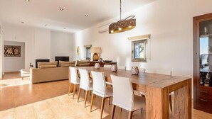 A spacious living area with a wooden dining table, white chairs, a sofa, and a television.