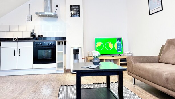 Cocina moderna con armarios blancos, encimera negra y horno empotrado. Se observa un televisor con pantalla verde sobre un soporte de madera, un sofá marrón y una mesita con un florero.