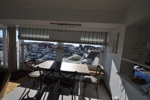 A dining area with a table and chairs, a view of houses through large windows, and a kitchen area with cabinets and a counter. - Apartment in Duinbergen With Sea Views (Knokke-Heist)