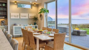 A dining area with a table set for a meal, wicker chairs, a white cabinet, and a view of a deck with a fire. - Chic Whidbey Island Waterfront Home w/ Hot Tub, Firepits + Views (Coupeville)