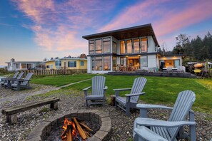 Une maison moderne avec un foyer extérieur et des fauteuils Adirondack dans le jardin.