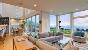 A spacious living area with a large sofa, a dining table, and a kitchen in the background. - Chic Whidbey Island Waterfront Home w/ Hot Tub, Firepits + Views (Coupeville)