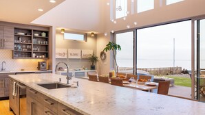 A modern kitchen with a large island, a dining area with wicker furniture, and a view of the sea. - Chic Whidbey Island Waterfront Home w/ Hot Tub, Firepits + Views (Coupeville)