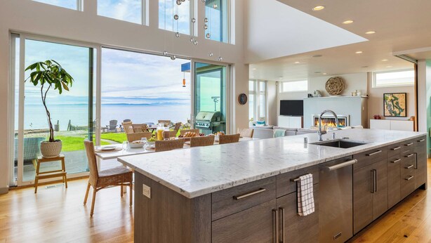 A modern kitchen with a large island, a dining area with outdoor views, and a cozy seating area with a fireplace. - Chic Whidbey Island Waterfront Home w/ Hot Tub, Firepits + Views (Coupeville)