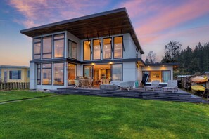 Une maison moderne à deux étages, dotée de grandes fenêtres, d’une terrasse et d’un espace salon extérieur.