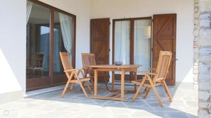 Un patio con mesa de madera y cuatro sillas, suelo de piedra y puertas de madera con paneles de vidrio.