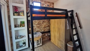 A bunk bed with a wooden ladder, a white shelving unit with various items, a small table with a patterned tablecloth, and a stone wall.