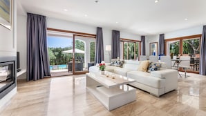 A modern living room with a white sofa, a coffee table, and a TV. There are large windows with curtains, a pool view, and a dining area with a table and chairs. - Villa Bella Vista in Sa Pobla (Sa Pobla)