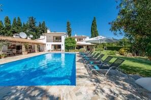 A swimming pool with lounge chairs and umbrellas, surrounded by a well-maintained garden and tall trees. - Villa Can Toni in Port de Pollen a (Pollenca)