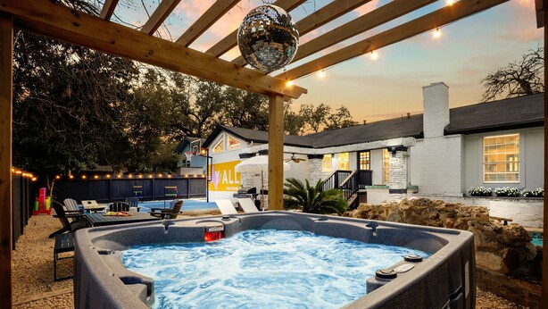 Un jacuzzi bajo una pérgola con luces colgantes, una piscina y una casa con un cartel que dice 'ALL'.