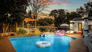 Una piscina en el patio trasero con anillos inflables, un pérgola con luces colgantes y un muro de piedra.