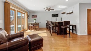 Un salón espacioso con un sofá de cuero, una zona de comedor con sillas de madera, un ventilador de techo y un televisor montado en la pared.