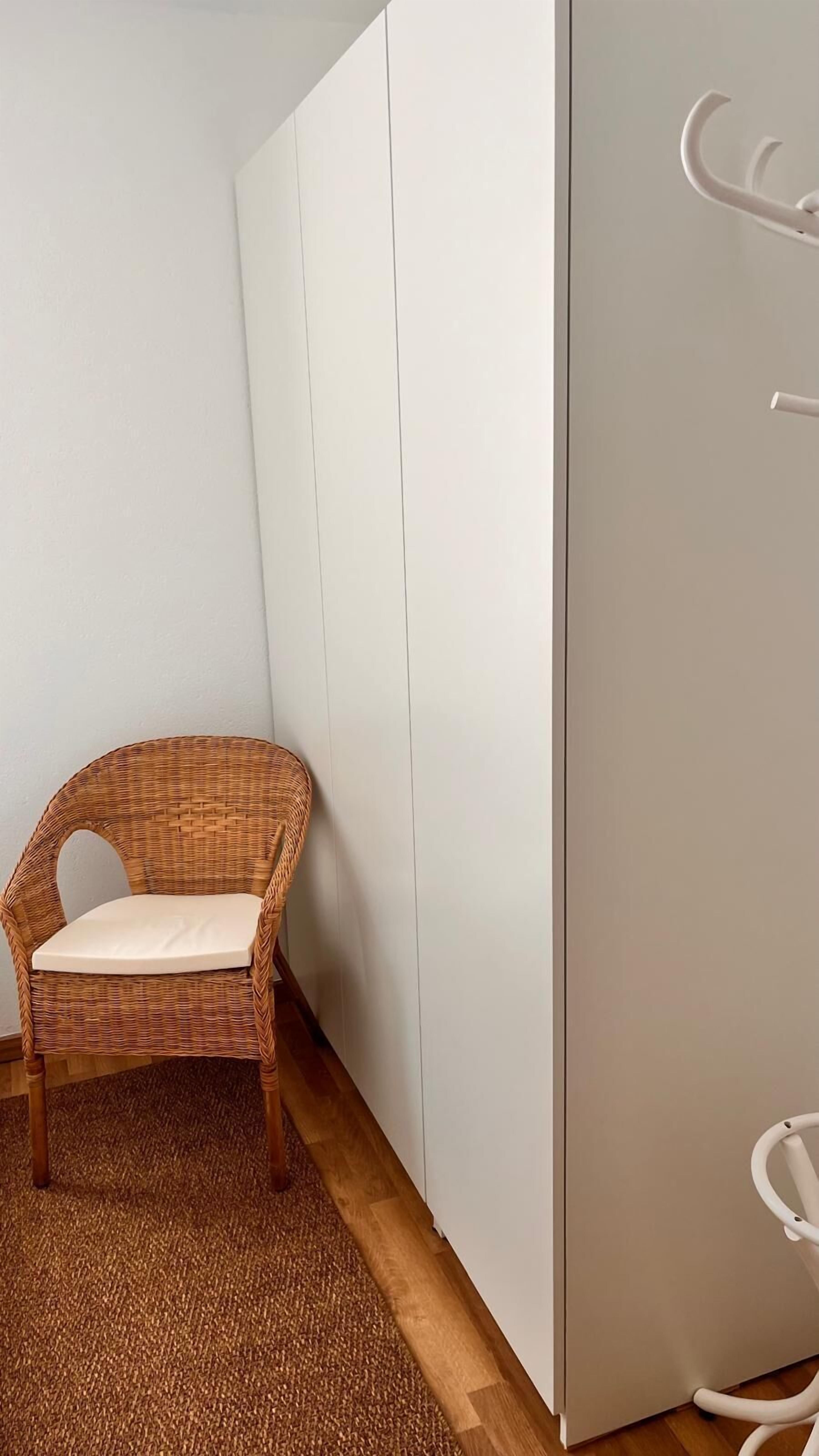 A room with a wicker chair, a white wardrobe, and a white cabinet.