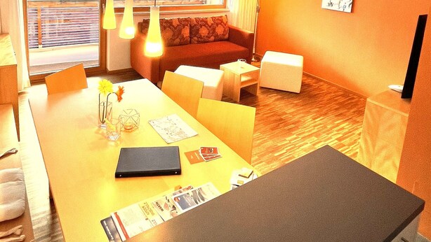 A modern hotel room with a large window offering a view of the ocean, a cozy seating area with a sofa and armchairs, a dining table with a vase of flowers, and a flat-screen TV mounted on the wall. - Adventure Panorama (Warth)