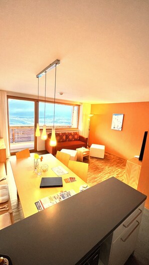 A modern hotel room with a large window offering a view of the ocean, a cozy seating area with a sofa and armchairs, a dining table with a vase of flowers, and a flat-screen TV mounted on the wall. - Adventure Panorama (Warth)