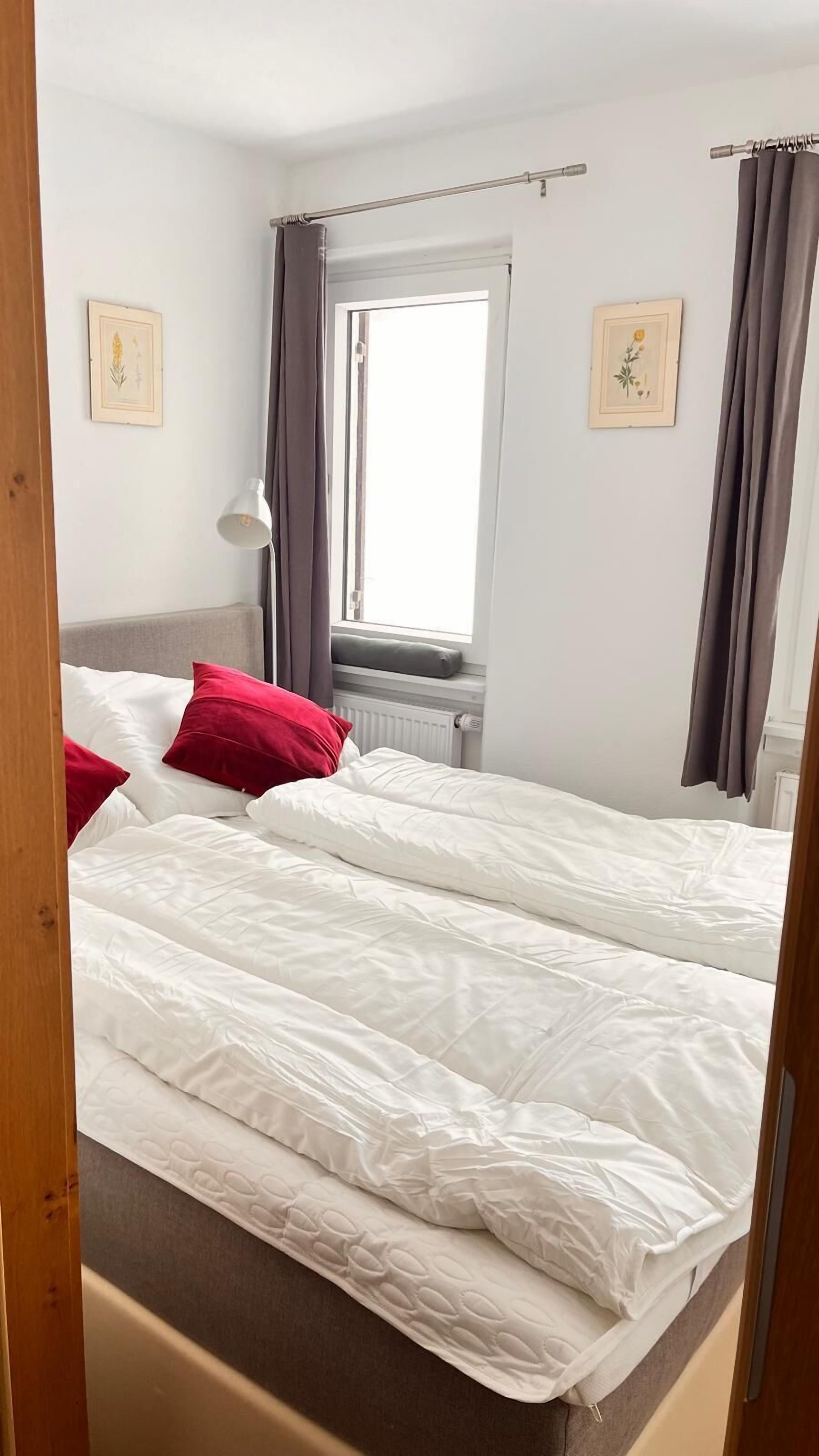 A double bed with white bedding and red pillows, a window with curtains, and two wall-mounted pictures.