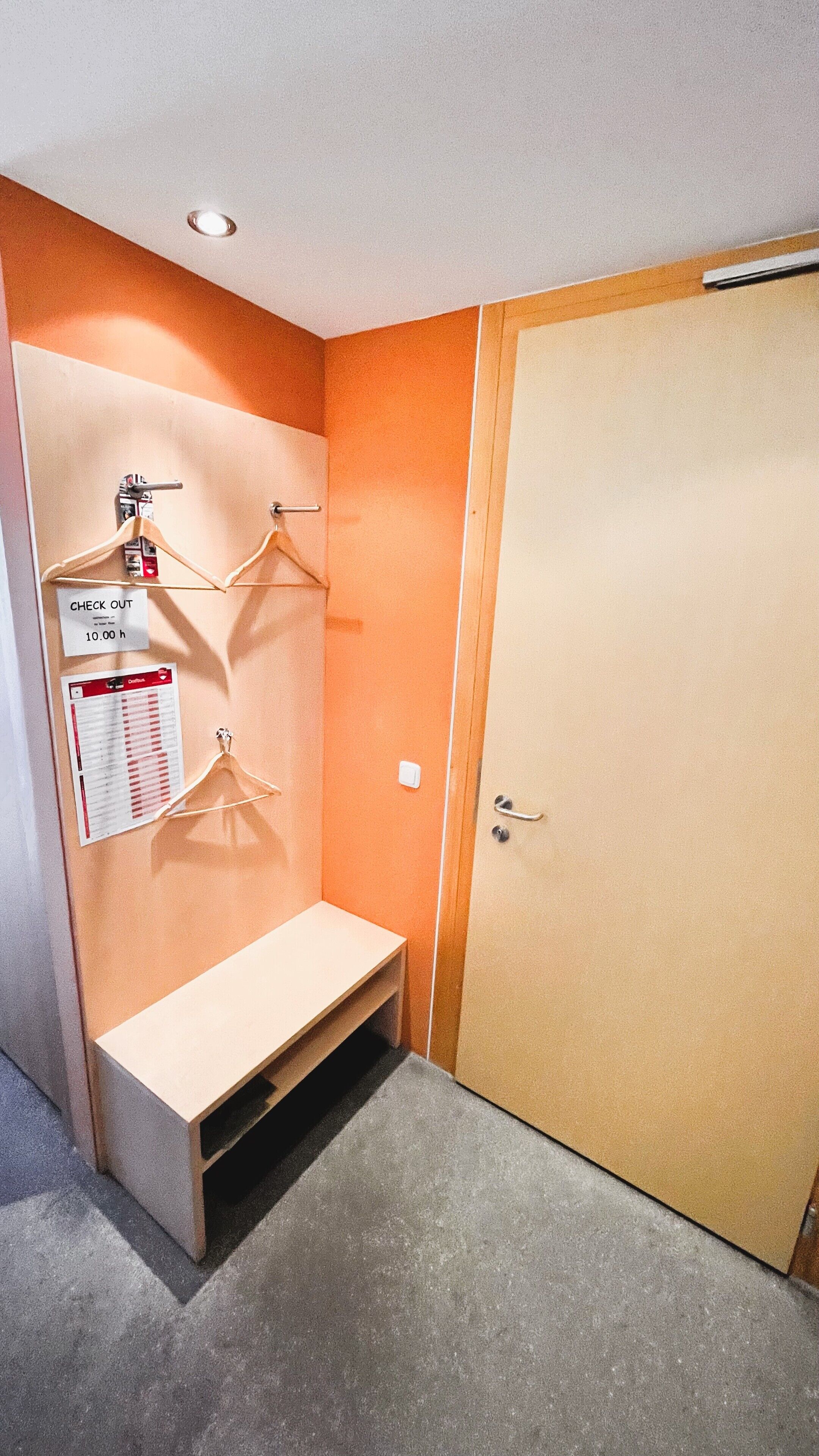 Ein kleiner, kompakter Raum mit einem Einzelbett, einem Holzschrank, einer Bank und einem an der Wand befestigten Kleiderhaken mit Haken. An der Wand befindet sich ein Schild mit der Aufschrift 'CHECK OUT' sowie eine Auflistung von Gegenständen.
