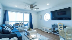A modern living room with a blue sofa, a coffee table, a TV mounted on the wall, and a ceiling fan.