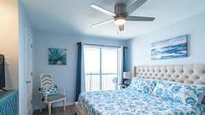 A bedroom with a bed, a chair, a ceiling fan, a window with curtains, and artwork on the wall.