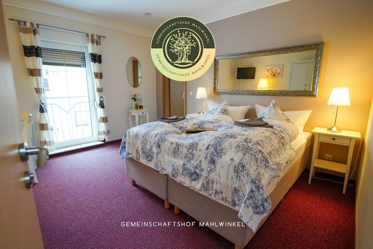 A hotel room with a bed, bedside tables, a mirror, a window with curtains, and a red carpet.