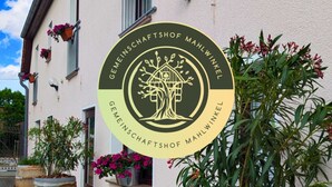 A building with a sign reading "zu fällig wien" and a logo for "GEMEINSCHAFTSHOF MAHLWINKEL". There are potted plants and a table with chairs outside. - Gemeinschaftshof Mahlwinkel (Angern)