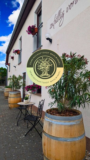 A building with a sign reading "zu fällig wien" and a logo for "GEMEINSCHAFTSHOF MAHLWINKEL". There are potted plants and a table with chairs outside. - Gemeinschaftshof Mahlwinkel (Angern)