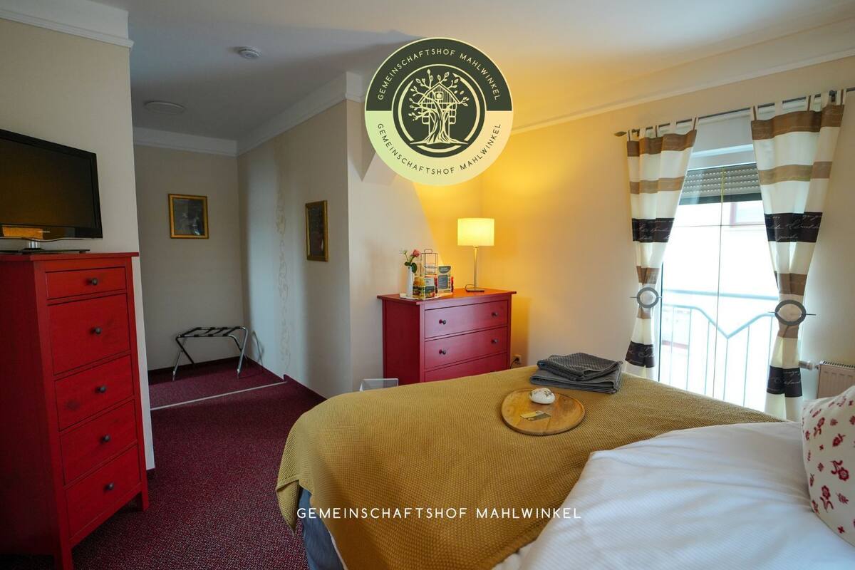 A hotel room with a bed, a red dresser, a television on top of the dresser, a lamp, and a window with curtains.