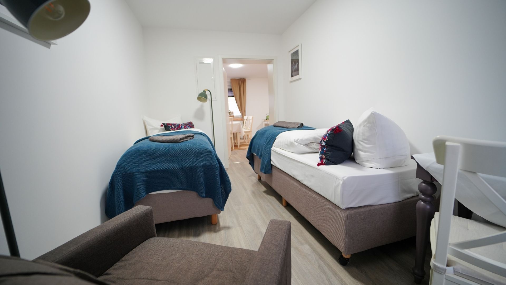 Habitación de hotel estrecha con dos camas, una silla y una mesita.