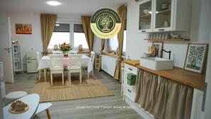 A modern kitchen with a dining area, featuring a white table, chairs, and a rug. There is a cabinet with glass doors, a small refrigerator, and a window with curtains.