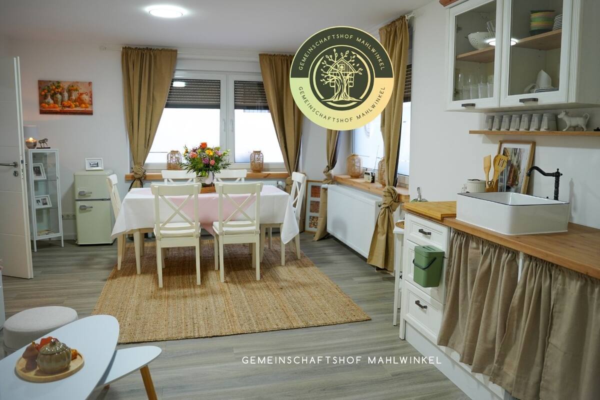A modern kitchen with a dining area, featuring a white table, chairs, and a rug. There is a cabinet with glass doors, a small refrigerator, and a window with curtains.