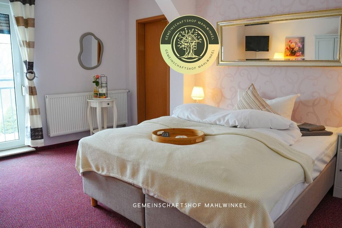 A hotel room with a bed, bedside tables, a mirror, a radiator, a window with curtains, and a wall with a logo.