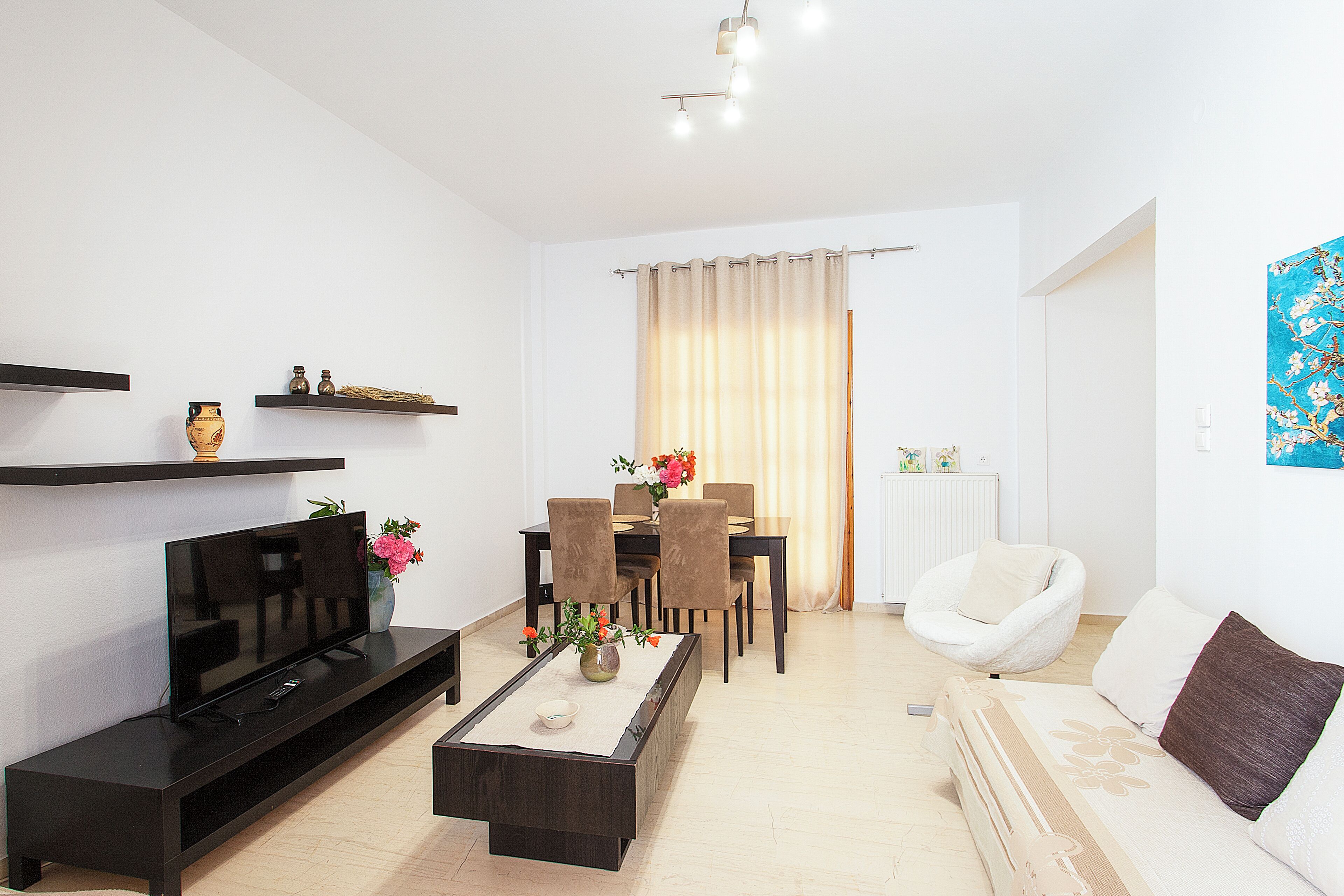 Un salon moderne avec un téléviseur à écran plat, une table basse ornée de fleurs, un coin repas avec des chaises et un canapé blanc.