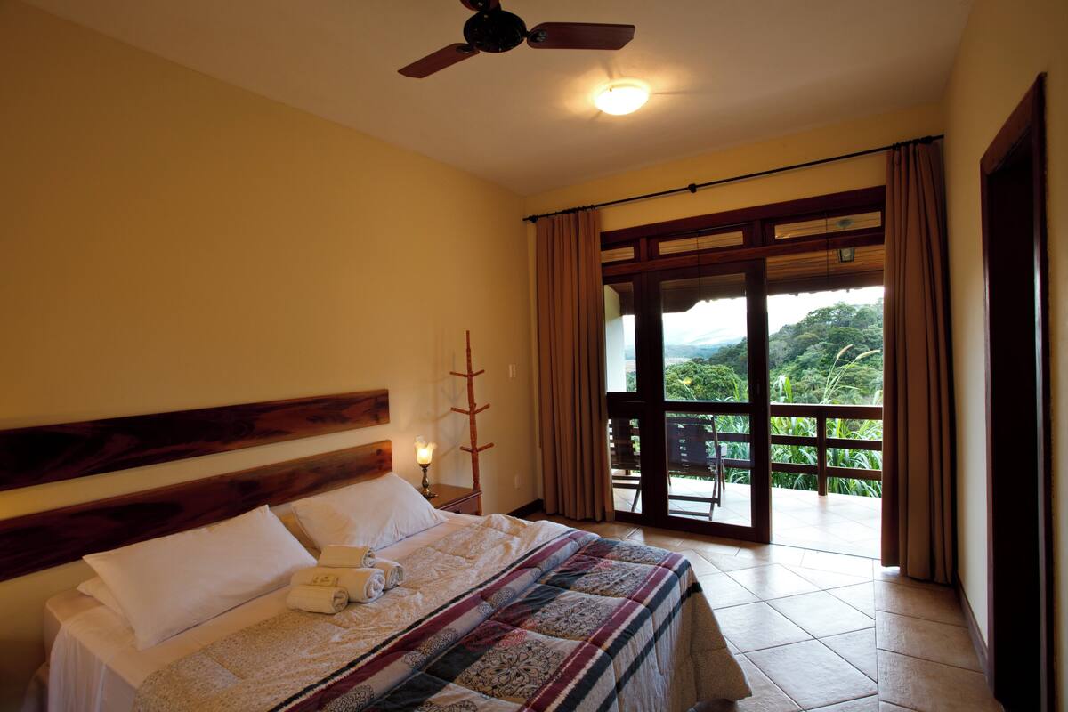 A hotel room with a bed, a ceiling fan, a nightstand with a lamp, and a view of the outdoors through a sliding door.